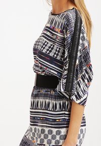 Sukienka drukowana z wielokolorowymi wzorami, luźny krój, szerokie rękawy i czarny pasek w talii. Materiał jest lekki i teksturowany.