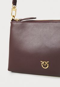 Burgundska kožna torba s glatkom teksturom, zlatnim metalnim detaljima, zatvaranjem na zip i dekorativnim logotipom s dva ptice sprijeda.