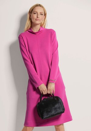 Roze lange mouw jurk met een cowl nek, met een losvallende pasvorm en zijzakken. Het model houdt een kleine zwarte handtas van textuurmateriaal vast.