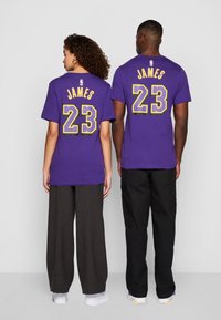 Fialové bavlněné trička s potiskem "JAMES" a "23" ve zlaté a černé na zadní straně, nošena s volnými kalhotami a bílými botami.
