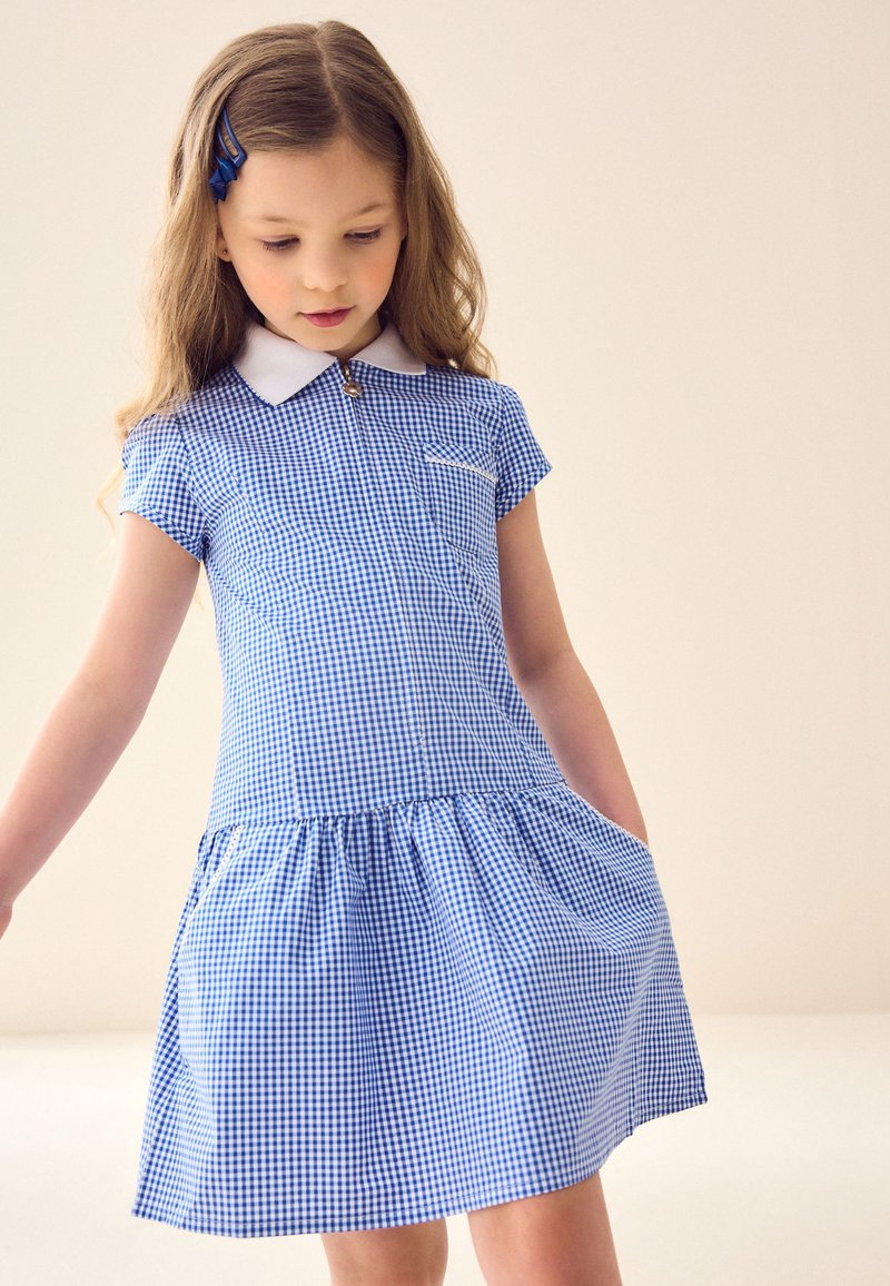 Junges Mädchen mit langen Haaren und einer blauen Haarspange, das ein blau-weiß kariertes kurzärmeliges Kleid mit weißem Kragen trägt und nach unten schaut.