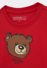 T-shirt en coton organique rouge avec un graphique de ours en dessin animé de couleur marron avec des yeux noirs. Texte "GUESS EST.1981" sous le design de l'ours.