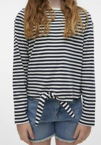 Camicia a maniche lunghe a righe in cotone navy e bianca con un dettaglio anteriore annodato, abbinata a pantaloncini di denim con orli sfrangiati.