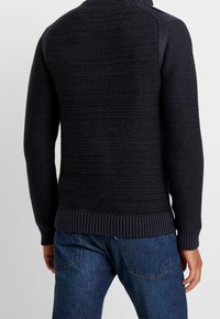 Mörkstickad tröja med ribbad nederkant och muddar. Texturerad, figurnära design med hög krage och horisontell randning på kroppen.