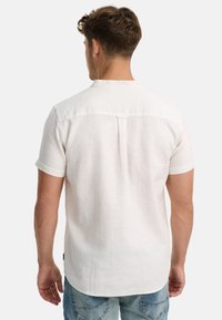 Man in een wit T-shirt met korte mouwen en een blauwe spijkerbroek, met zijn rug naar de camera gekeerd, staande tegen een effen lichte achtergrond.