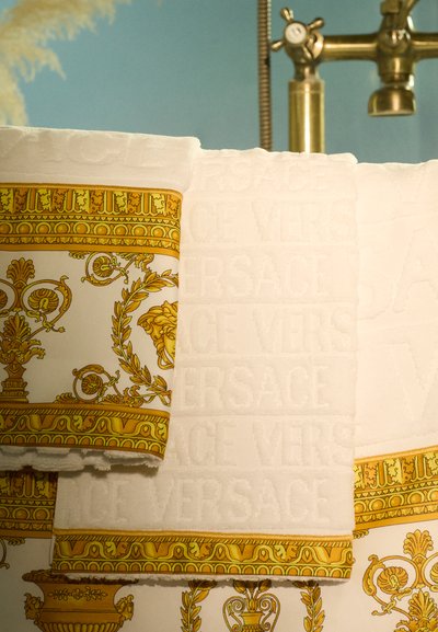 Des serviettes blanches et dorées présentent des motifs complexes et une texture raffinée, avec "VERSACE" estampé sur le tissu et une bordure décorative.