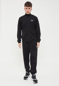 Tuta nera realizzata in un tessuto morbido ed elasticizzato. Presenta una giacca con zip e colletto alto, polsini elasticizzati e pantaloni coordinati con accenti del logo.