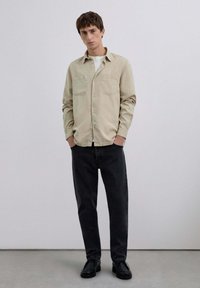Camisa beige de botón con dos bolsillos en el pecho, combinada con jeans negros y zapatos negros. Tejido suave, ajuste estructurado y un diseño sutil.