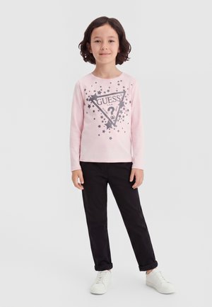 Barn med kort mørkt hår iført en pink langærmet trøje med stjerner og sorte bukser, står på en ensfarvet lys baggrund.