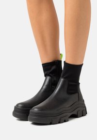 Svarta anklarstövlar med en slät läderkropp, elastisk tygövre, djärv klumpig sula och neongrön dragrem för enkel påtagning.