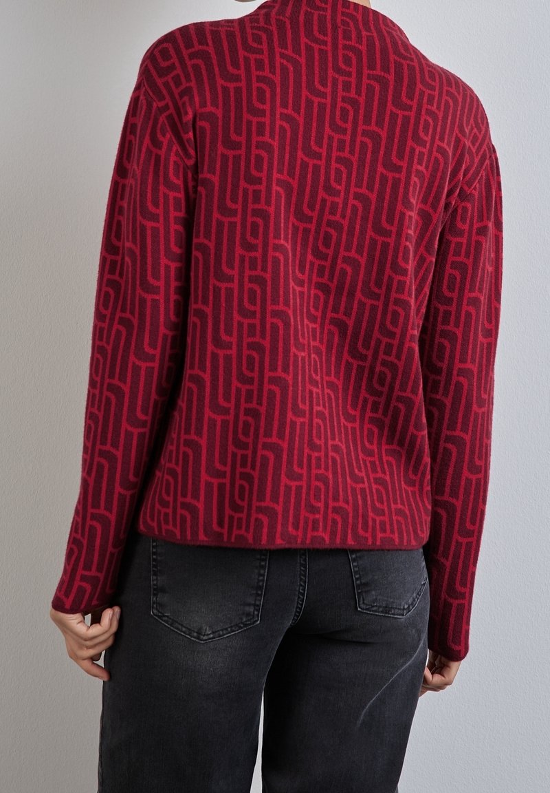 Persoon die een rood shirt met lange mouwen draagt met een donker geometrisch patroon en zwarte spijkerbroek, gezien van achteren tegen een effen muur.