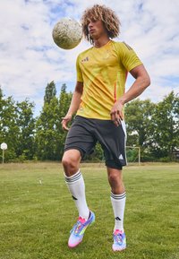 Gul atletik trøje med røde detaljer, sorte shorts, hvide knæhøje sokker og multicolor fodboldstøvler. Spiller jonglerer med en fodbold.