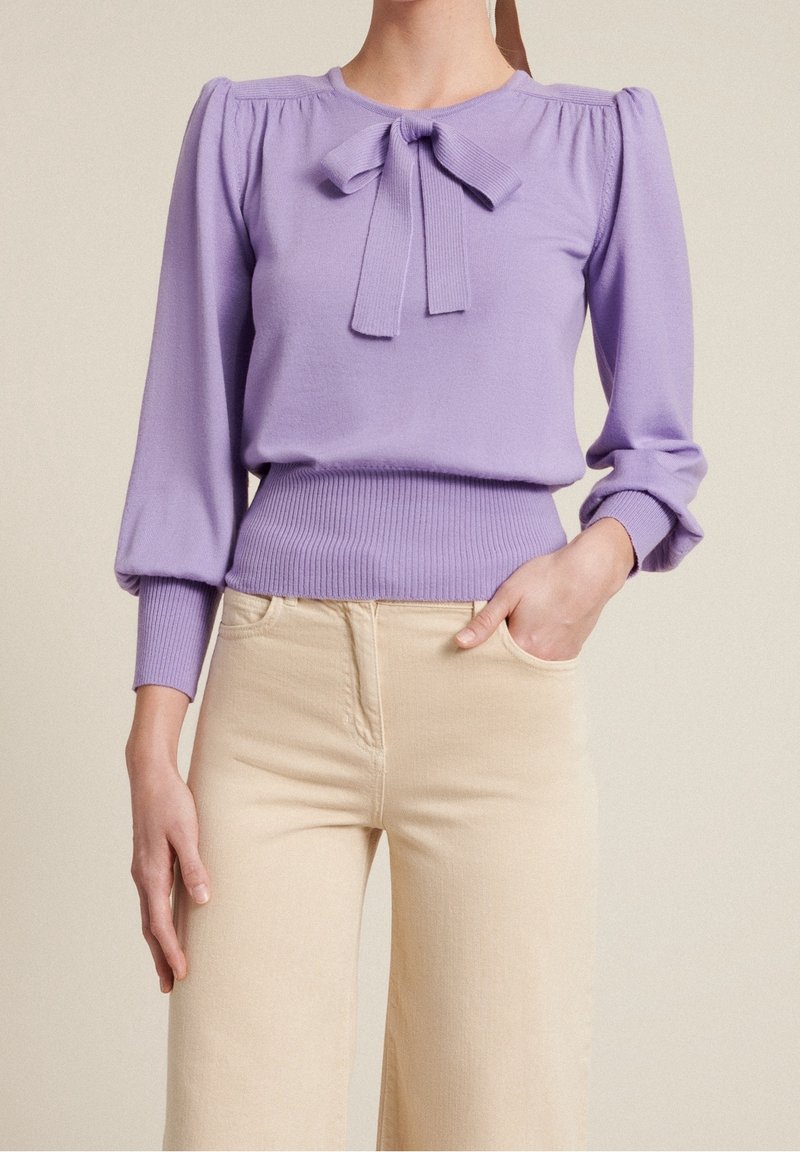 Lila gebreide trui met pofmouwen en een strikdetail bij de hals. Gecombineerd met beige broek met wijde pijpen en een aansluitende tailleband.