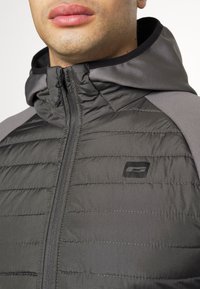 Svart pufferjacka med huva, med quiltad textur och elastiska detaljer. Dragkedjestängning och en logotyp-patch på bröstet.