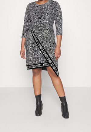 Femme portant une robe portefeuille mi-longue à motifs abstraits noirs et blancs, manches trois-quarts, et des bottines noires.