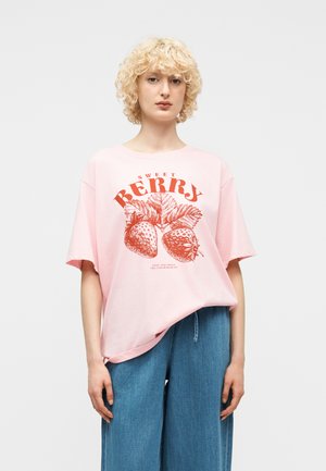 Person med krøllet blondt hår iført en pink oversized T-shirt med røde jordbærmotiver og blå bredbenede jeans.