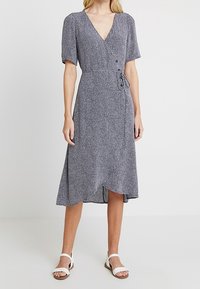 Navyblaues Wickelkleid mit weißen Punkten; verfügt über kurze Puffärmel, einen V-Ausschnitt und eine gebundene Taille, die bis zur Mitte der Wade reicht. Weiße Sandalen.