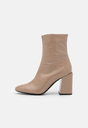 Beige ankelstøvle lavet af glat læder, med spids tå og en chunky blokhæl med metallic-accenter. Lynlås i siden.