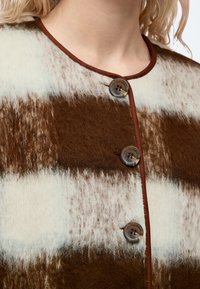 Gros plan d’une personne portant une veste rayée marron et blanche en tissu duveteux avec des boutons ronds et une bordure marron autour du col.