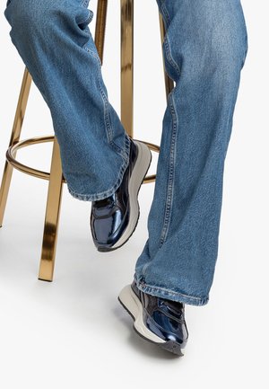 Persona seduta su uno sgabello di metallo dorato, indossando jeans blu e sneakers scure e lucide, con uno sfondo bianco.