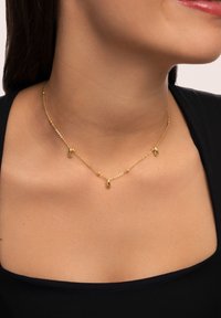 Collier en or avec une chaîne délicate ornée de trois pendentifs en forme de goutte, maillons texturés et finition polie, porté sur une peau nue.