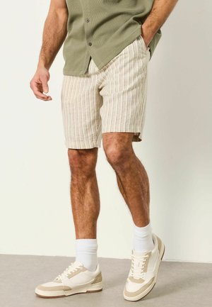 Man wearing beige striped shorts, white socks, beige and white sneakers, and an olive green short-sleeve shirt with hand in pocket.