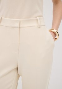 Beige structured trousers featuring a textured fabric, front pockets, and a belt loop waistband. Accompanied by a gold circular bangle.