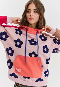 Ružová mikina s veľkými námorníckymi kvetinovými vzormi, koralovou kapucňou, modrými šnúrkami a koralovou prednou vreckom s nálepkou. Mäkká textúra látky.