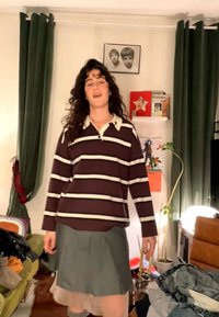 Γυναίκα με σκουρόχρωμα κατσαρά μαλλιά, που φοράει μία ριγέ μακρυμάνικη μπλούζα και γκρι φούστα, στέκεται σε ένα ακατάστατο σαλόνι με πράσινες κουρτίνες και έργα τέχνης στον τοίχο.