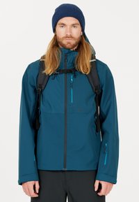 Teal vattentät jacka med huva, framdragkedja och bröstficka; justerbara ärmmuddar och två sidofickor. Buren med en svart ryggsäck.