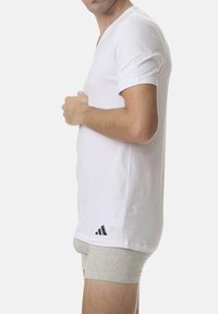 White short-sleeve V-neck t-shirt made of cotton, featuring a black Adidas logo near the hem. Model is wearing grey shorts.