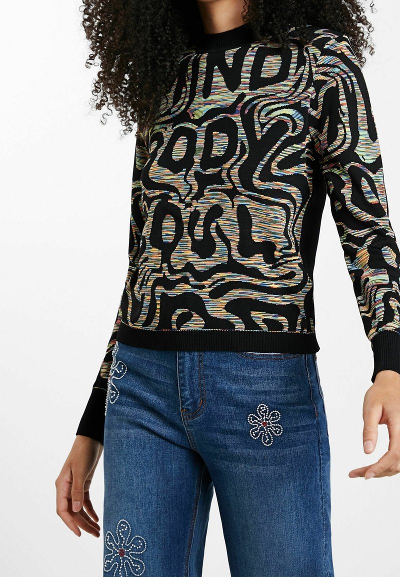 Femme portant un pull noir avec un motif abstrait ondulé multicolore et des lettres épaisses, associé à un jean bleu orné de perles florales.
