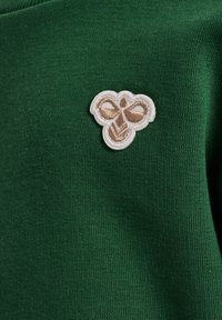 Grön bomullstyg har en liten broderad bi-logotyp i beige, omramad i vitt, placerad på den övre vänstra sidan.