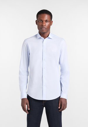 Junger Mann mit dunkler Hautfarbe, der ein hellblaues, langärmliges Hemd mit Knöpfen und dunkle Jeans trägt und vor einem schlichten weißen Hintergrund steht.