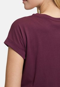 Burgunder T-Shirt mit kurzen aufgerollten Ärmeln, aus weichem Baumwollmaterial und mit lässiger Passform; sichtbare Nähte entlang des Halsausschnitts und der Ärmel.