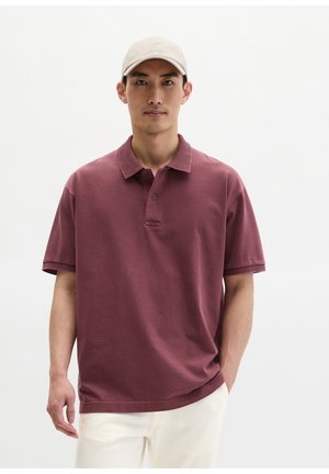 Mann trägt eine beige Mütze und ein kastanienbraunes Polo mit kurzen Ärmeln sowie eine weiße Hose, steht vor einem einfarbigen hellen Hintergrund.