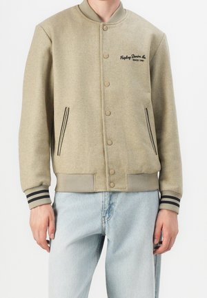 Beżowa kurtka varsity z prążkowanym kołnierzem i mankietami z czarnymi paskami, zapinana na guziki, z bocznymi kieszeniami, haftowanym logo "Replay Denim Co. Since 1981".