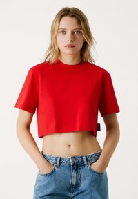 Top crop rosso realizzato in tessuto solido, con colletto rotondo e maniche corte. Indossato con jeans in denim leggero a vita alta di colore blu.