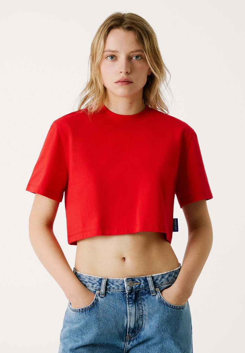Top crop rosso realizzato in tessuto solido, con colletto rotondo e maniche corte. Indossato con jeans in denim leggero a vita alta di colore blu.