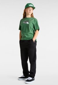 Grünes Baumwoll-T-Shirt mit Grafik auf der Brust, schwarze Hose und schwarze Vans-Schuhe mit weißen Sohlen. Grüne Kappe mit dem "VANS"-Logo.