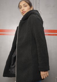 Manteau en fausse fourrure noire avec capuche, doté d'une fermeture éclair et de deux poches latérales. La texture est douce et duveteuse, offrant chaleur et confort.