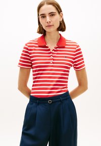 Γυναίκα που φοράει κόκκινο και λευκό ριγέ πόλο πουκάμισο, μπλουζαρισμένο σε ψηλόμεσο σκούρο μπλε παντελόνι με λεπτή μαύρη ζώνη, στέκεται με τα χέρια πίσω από την πλάτη.