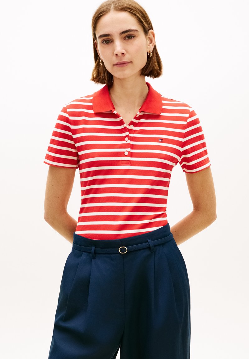 Γυναίκα που φοράει κόκκινο και λευκό ριγέ πόλο πουκάμισο, μπλουζαρισμένο σε ψηλόμεσο σκούρο μπλε παντελόνι με λεπτή μαύρη ζώνη, στέκεται με τα χέρια πίσω από την πλάτη.