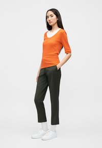 Πορτοκαλί εφαρμοστό πουκάμισο με κοντά μανίκια, σε συνδυασμό με σκούρους πράσινους παντελόνες slim-fit. Το μοντέλο φοράει λευκά αθλητικά παπούτσια και λευκές κάλτσες μέχρι τον αστράγαλο.