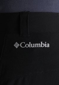 Tissu noir avec le logo blanc de la marque Columbia imprimé au-dessus d’une passants de ceinture sur un pantalon ou un short.