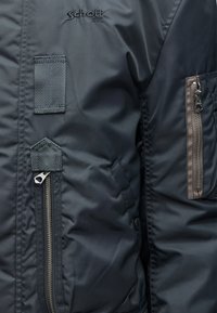 Gros plan d'une veste bleu foncé avec deux poches verticales zippées et un logo Schott NYC brodé sur la poitrine.