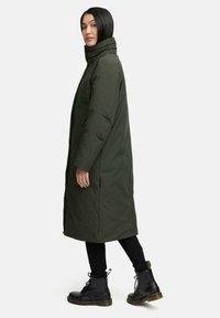 Langer grüner Daunenmantel mit hohem Kragen, Seitentaschen und Nähten. Getragen über schwarzen Leggings und schwarzen Schnürstiefeln.