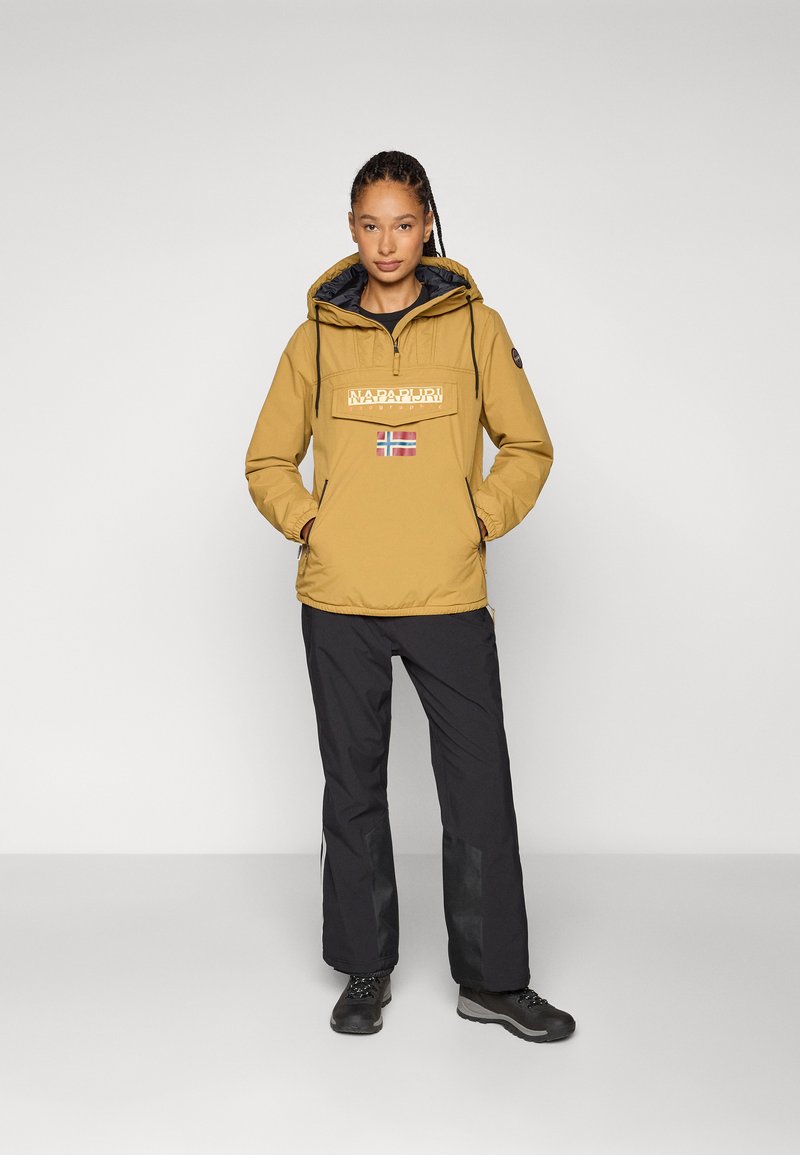 Gelber Pullover-Jacke mit Kordelzugkapuze, Frontlogo und norwegischem Flaggenmotiv; kombiniert mit schwarzen Hosen und dunklem Schuhwerk.