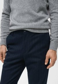 Szary dzianinowy sweter z prążkowanymi mankietami w zestawieniu z ciemnogranatowymi spodniami w cienkie prążki. Zbliżenie na dolną część tułowia, ukazujące strukturę tkaniny i dopasowanie.