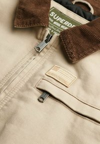 Veste beige avec col en velours côtelé marron, fermeture éclair en métal et poche avant arborant un patch logo Superdry avec design de drapeau.